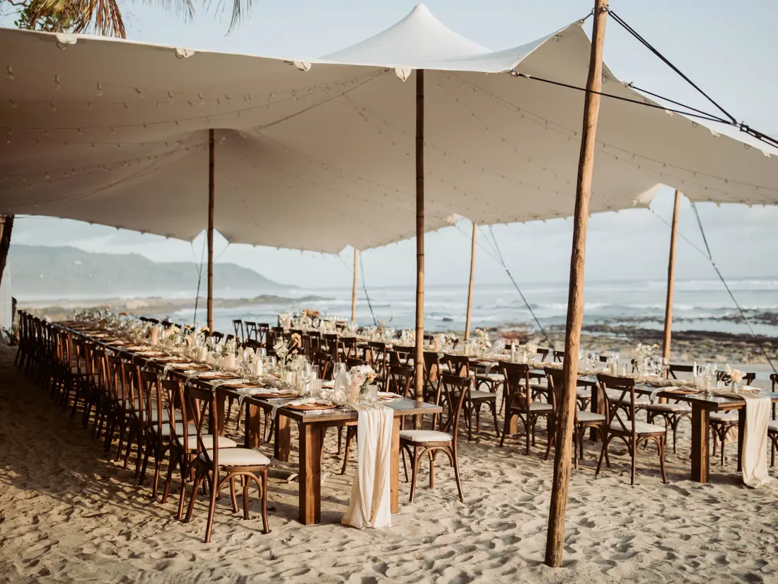 Long banquet tables set on the sand beneath a stretch tent hung with string lights, the surf breaking behind