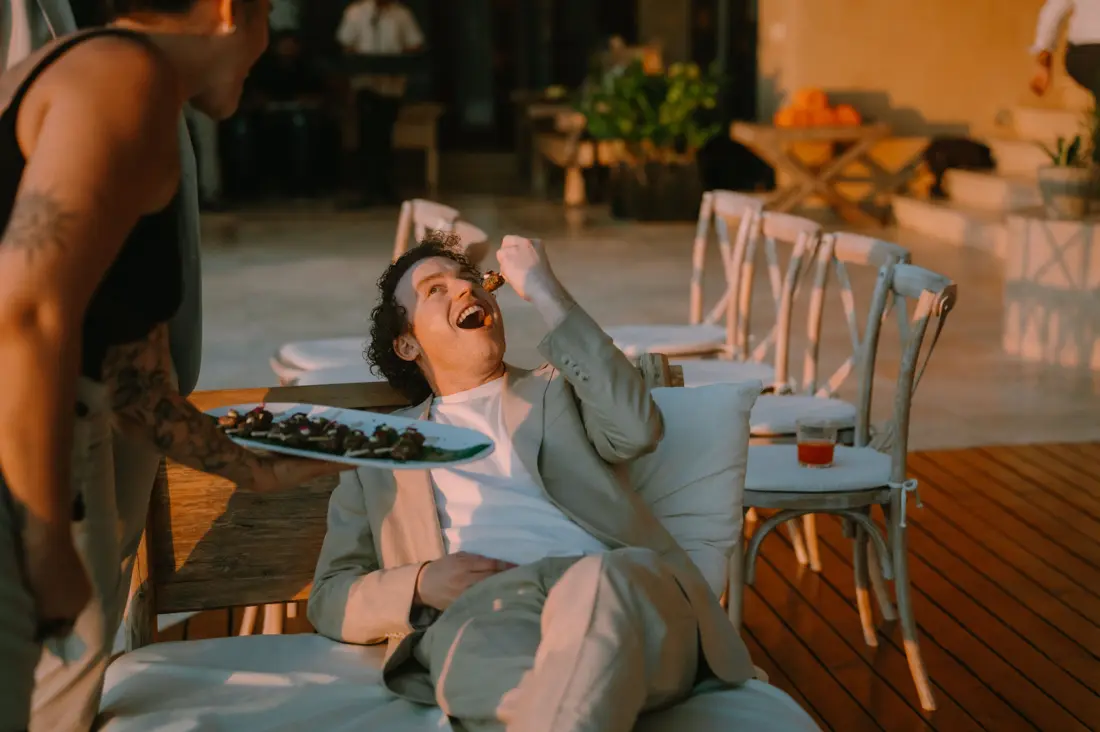 A guest stretched out on a daybed at golden hour, laughing as he takes a canapé from a platter held out beside him