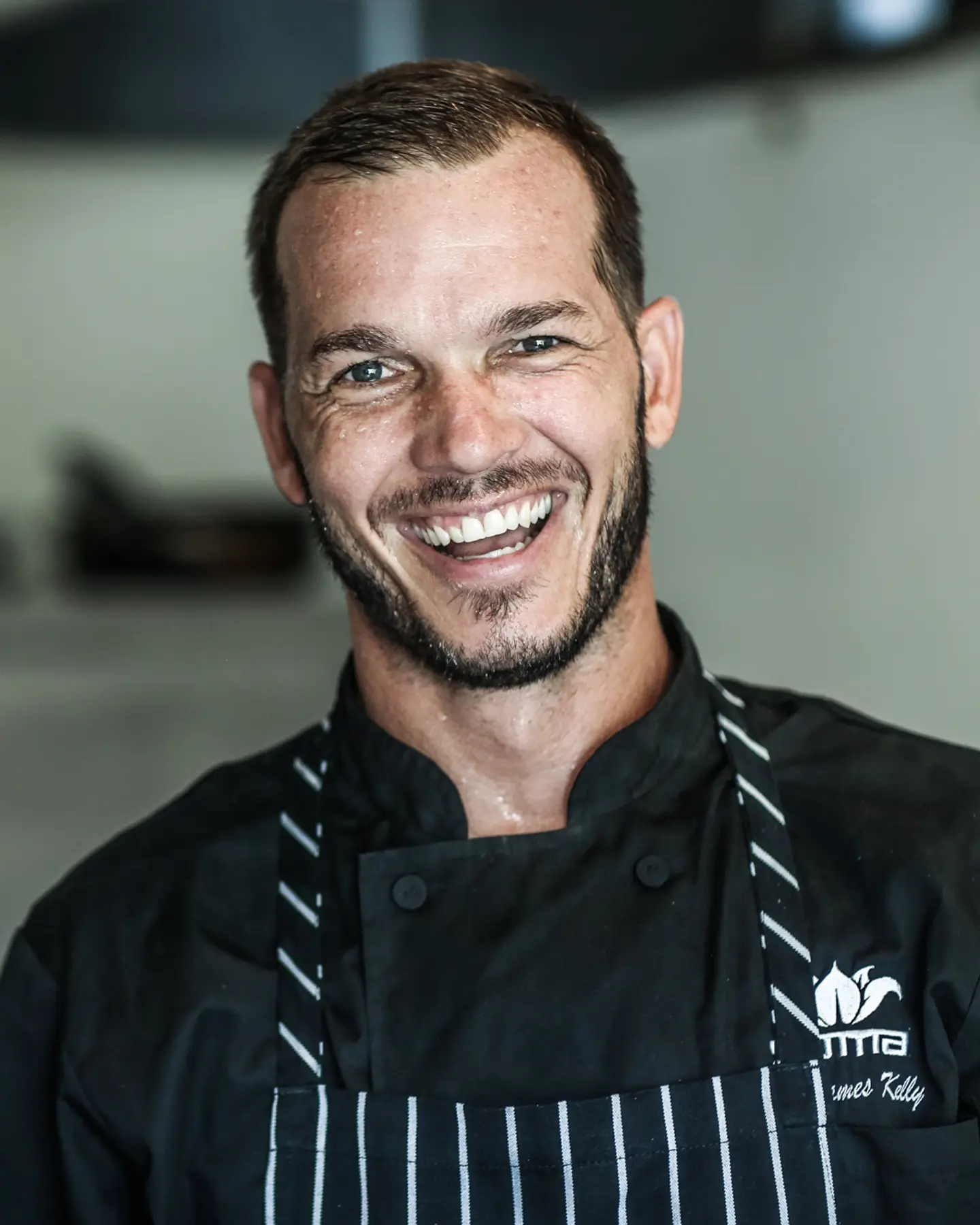 Chef James Kelly laughing in a stainless-steel kitchen, wearing a SOMA chef’s jacket and pinstriped apron