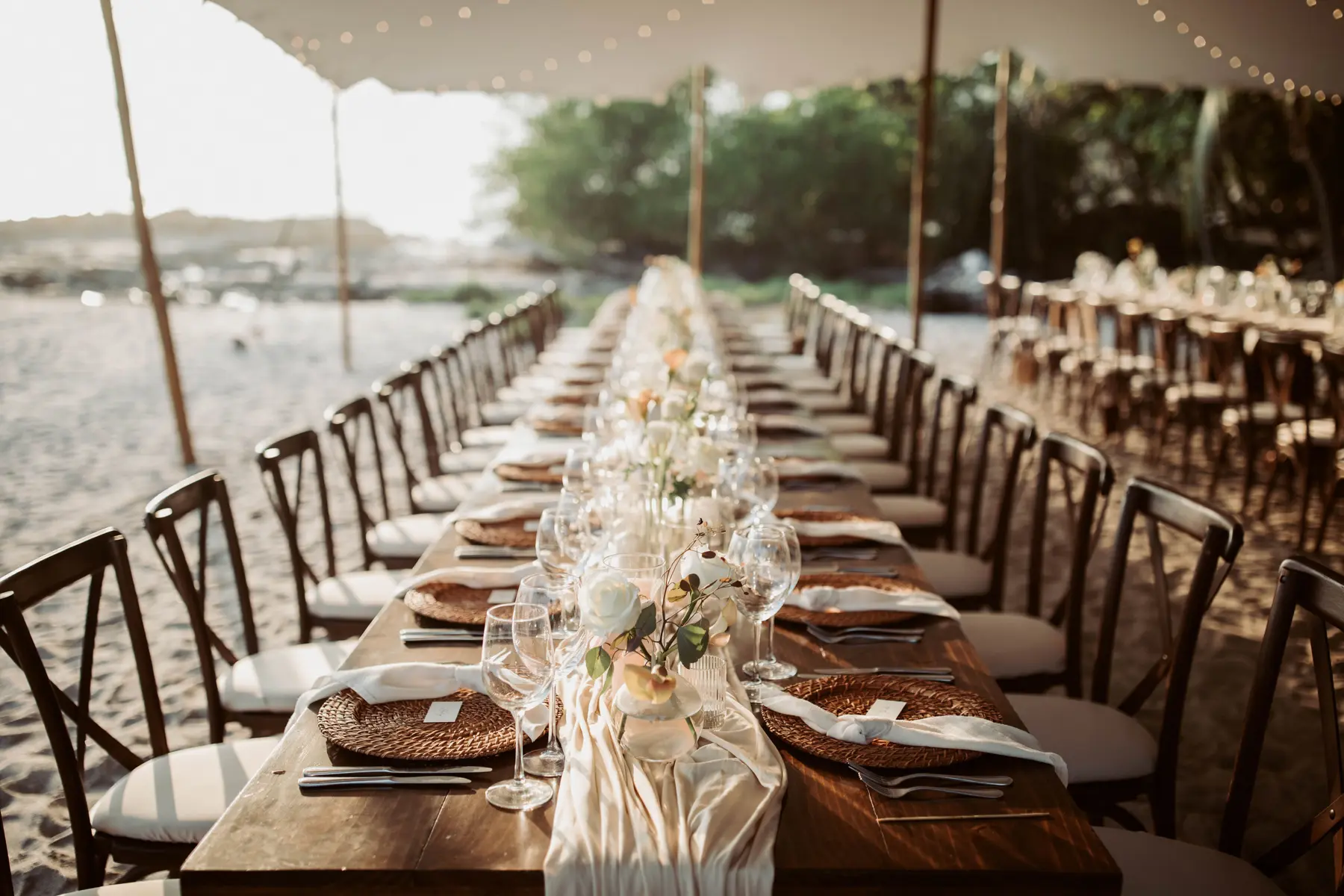 A long table laid on the sand under a stretch tent — woven chargers, linen runners and low garden roses receding down the table