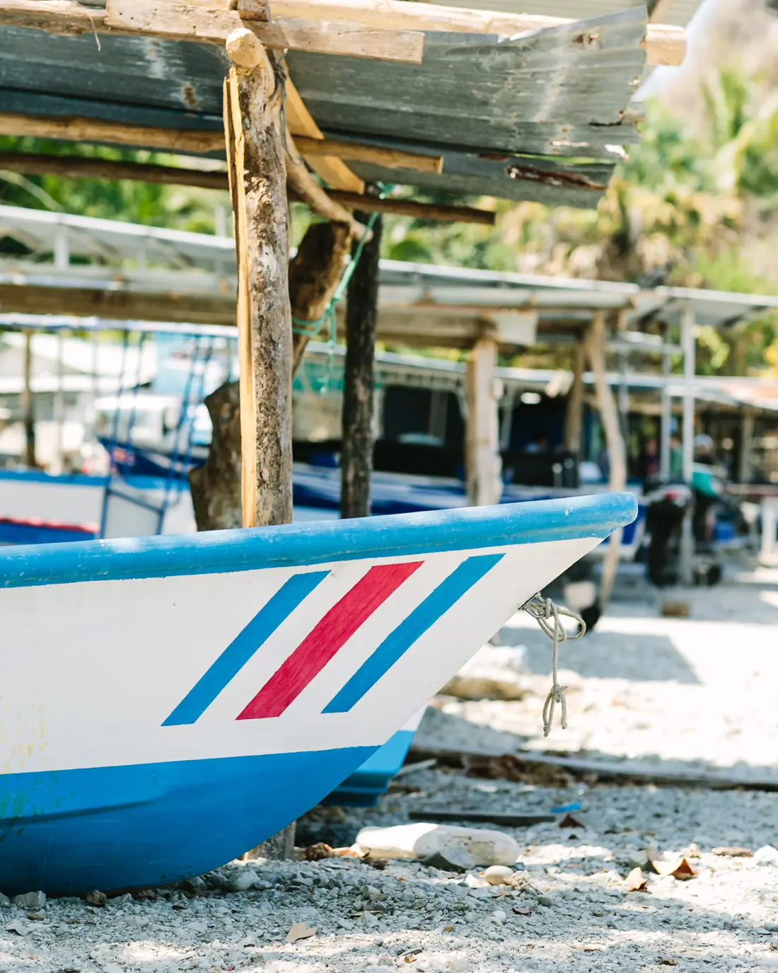 The painted bow of a fishing panga drawn up on a pebble beach beneath palapa shade