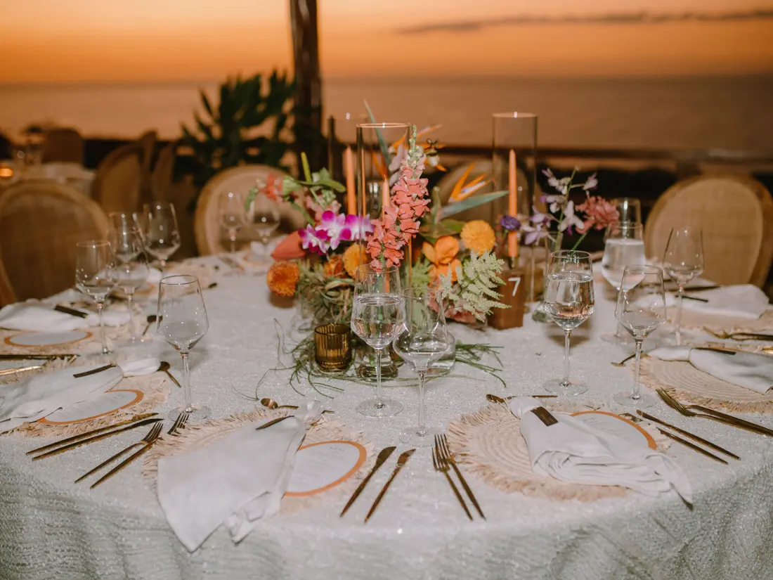 Table laid for dinner above the ocean at sunset — tapered candles, tropical florals and cane-backed chairs